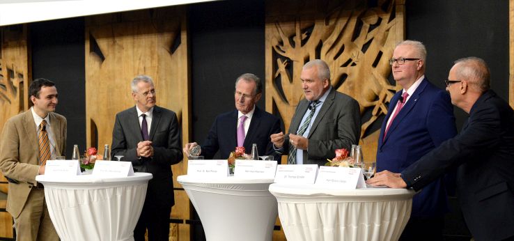 Die 5 Podiumsdiskussionsteilnehmer plus der Moderator stehen auf der Bühne der Aula um Stehtische gruppiert (Bild: ivwKöln / TH Köln / Gerhard Richter)