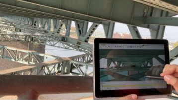 Ein Tablet mit einem 3D-Modell einer Brücke. Im Hintergrund die Tragkonstruktion der Brücke