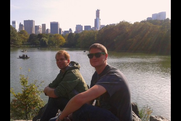 Zwei junge Männer sitzen vor einem See, im Hintergrund die Skyline von New York