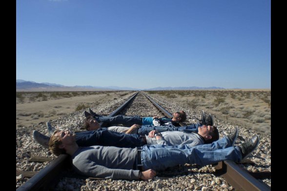 Vier junge Männer liegen quer über ein Gleis in endloser Prärie