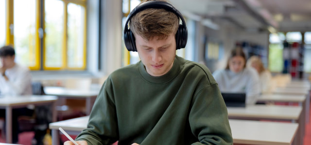 Ein männlich gelesener Studierender arbeitet mit Kopfhörern, einem iPad und einem Buch an einem Tisch. Im Hintergrund sitzen weitere Studierende (Bild:Hanna Witte/TH Köln)