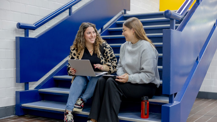 Zwei weiblich gelesene Studierende sitzen mit einem Laptop und einem iPad auf einer Treppe