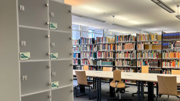 Am linken Bildrand sind Dauerschließfächer zu sehen. Rechts im Bild sind Stühle und Tische mit Bücherregalen im Hintergrund zu sehen. (Bild: TH Köln/ Alina Beier)