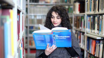 Studientin stehtmit einem geöffneten Buch zwischen Bücherregalen in der Bibliothek (Bild: FH Köln/Thilo Schmülgen)