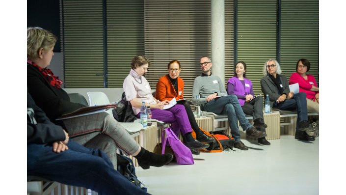 Bernadette Drescher beugt sich sitzend vor und spricht Claudia Middendorf an, die ganz links auf dem Bild zu sehen ist