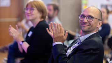 Ein Mann schaut lächelt in die Kamera und klatscht. Im Hintergrund applaudieren weitere Menschen. (Bild: Thilo Schmülgen/TH Köln)