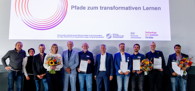 Elf Personen stehen auf der Bühne, einige halten einen Blumenstrauß oder einen Preis. Über ihnen eine Präsentation: 
