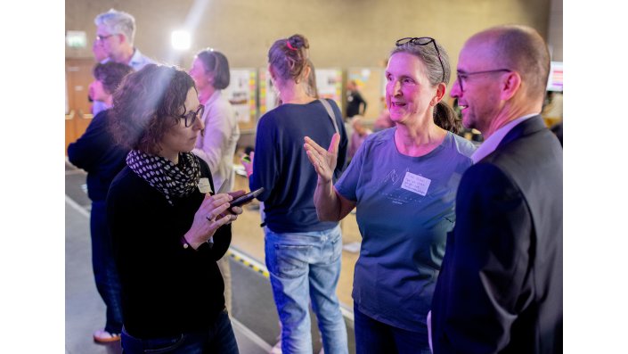 Drei Personen unterhalten sich vor einer Reihe von Pinnwänden mit Postern der Lehr- und Studienpreis-Einreichungen. Im Hintergrund betrachten weitere Personen die Poster.