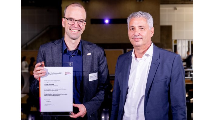 Zwei Personen stehen auf der Bühne, eine hält einen Preis.