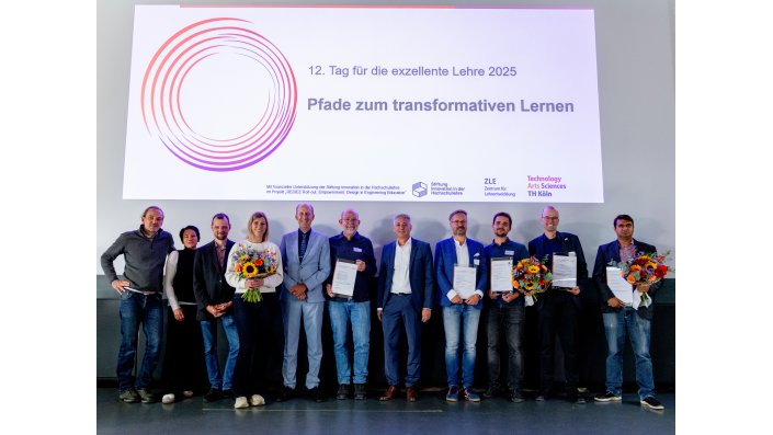 Elf Personen stehen auf der Bühne, einige halten einen Blumenstrauß oder einen Preis. Über ihnen eine Präsentation: "12. Tag für die exzellente Lehre: Pfade zum transformativen Lernen"