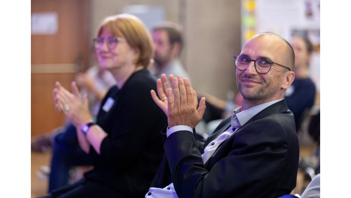 Ein Mann schaut lächelt in die Kamera und klatscht. Im Hintergrund applaudieren weitere Menschen.