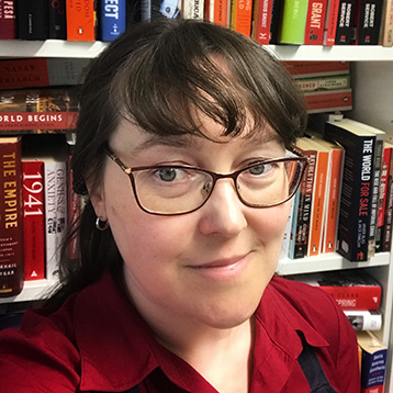 [Foto] Person mit braunen Haaren, Brille und rotem Hemd vor einem Bücherregal.