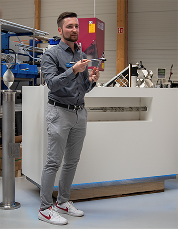 [Foto] Ein Mann in grauer Jacke und Jeans steht in einem Raum mit weißer Wand und Holzfußboden. Er hält ein Tablet in der Hand und trägt weiße Sportschuhe. Im Hintergrund sind Regale mit Ordnern, ein Schreibtisch mit einem Laptop und ein weißer Kühlschrank zu sehen.