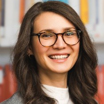 [Foto] Person mit langen, gewellten braunen Haaren, trägt eine Brille und eine graue Jacke über einem weißen Oberteil, unscharfer Hintergrund.