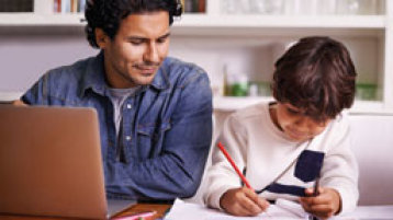 Ein junger Mann sitzt am Tisch, vor ihm ein aufgeschlagenes Laptop, neben ihm sitzt ein kleiner Junge und malt (Bild: iStock)