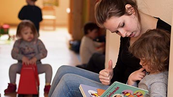 Eine Frau sitz neben einem Kind auf einer Bank und hält ein aufgeschlagenes Buch in der Hand. Im Hintergrund siueht man ein Kind auf einem BobbyCar. (Bild: Michaela Patschurkowski/TH Köln)