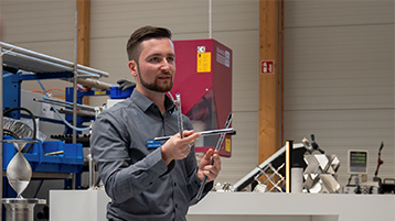 [Foto] Ein Mann in grauer Jacke und Jeans steht in einem Raum mit weißer Wand und Holzfußboden. Er hält ein Tablet in der Hand und trägt weiße Sportschuhe. Im Hintergrund sind Regale mit Ordnern, ein Schreibtisch mit einem Laptop und ein weißer Kühlschrank zu sehen. (Bild: privat)