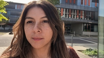 [Foto] Person mit langen braunen Haaren vor einem modernen mehrstöckigen Gebäude mit großen Fenstern und orangefarbenen Akzenten. (Bild: privat)