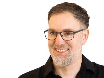 Jürgen Sleegers trägt ein schwarzes Hemd, eine schwarze Brille und dunkelbraune Haare. Er befindet sich vor einem weißen Hintergrund.