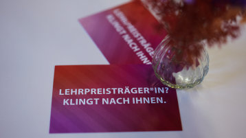 Zwei rote Postkarten mit weißer Schrift, die den Text 'LEHRPREISTRÄGER*IN? KLINGT NACH IHNEN.' tragen, daneben steht eine Vase mit Trockenblumen.  (Bild: ZLE TH Köln)