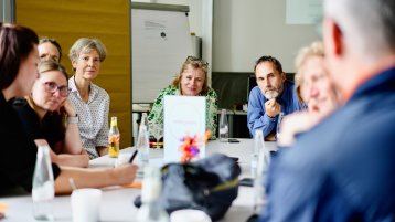 Acht Frauen und Männer sitzen an einem weißen Tisch mit Getränken und Kaffeetassen. Auf dem Tisch steht ein Schild mit dem Wort ,,arrangieren“. Im Hintergrund sieht man eine Leinwand mit einer Präsentation, ein Flipchart und eine Stellwand mit beigem Papier.  (Bild: ZLE TH Köln)