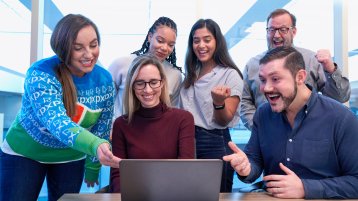 Vordergrund Laptop von hinten auf brauner Tischplatte. Hintergrund links 4 Frauen, rechts 2 Männer in einer Gruppe. Sie zeigen freudig und lachend auf den Laptop. (Bild: Windows / Unsplash)