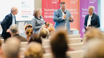 Foto: Drei Männer und eine Frau stehen vor einem Publikum und sprechen mit Mikrofonen, im Hintergrund sind Rollups mit der Aufschrift 'Technology Arts Sciences TH Köln' zu sehen. (Bild: Bild: Costa Belibasakis / TH Köln)