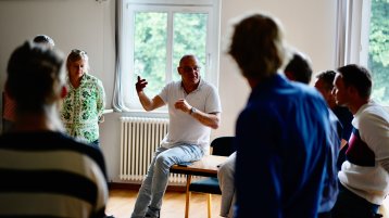In der Mitte sitzt Prof. Kreiser auf der Kante eines Tisches und rechts neben ihm steht Susanne Glaeser. Um ihn herum stehen und sitzen sechs Menschen in unscharfer Auflösung.  (Bild: ZLE TH Köln)