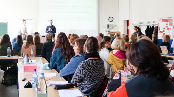 Die Mitglieder des EZ sitzen an langen Tischreihen. Im Hintergrund eine Präsentation (nicht lesbar) und zwei Sprecher. (Bild: ZLE / TH Köln)