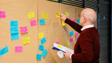 Herr Jörg Luderich steht vor der sandfarbenen Leinwand auf der gelb, rosa und hellblaue Post-Its hängen und er klebt ein gelbes mit schwarzer Schrift.