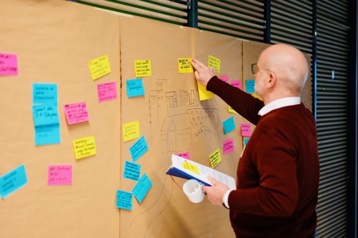 Herr Jörg Luderich steht vor der sandfarbenen Leinwand auf der gelb, rosa und hellblaue Post-Its hängen und er klebt ein gelbes mit schwarzer Schrift.