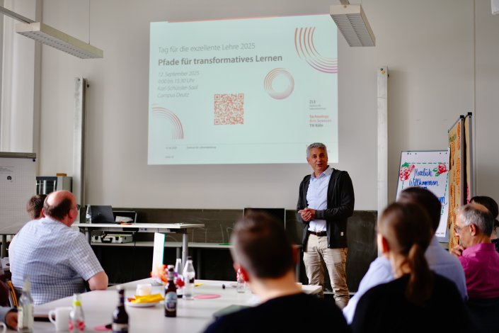 Rechts hält Prof. Dr. Axel Faßbender einen Vortrag. Hinter ihm wird die PowerPoint Präsentation auf einer Leinwand mit dem Titel ,,Tag für die exzellente Lehre 2025, Pfade für transformatives Lernen 12. September 2025. 9h bis 15 Uhr Karl-Schüssler-Saal Campus Deutz“ gezeigt. Daneben ist ein QR-Code abgebildet. Rechts hält Prof. Dr. Axel Faßbender einen Vortrag. Hinter ihm wird die PowerPoint Präsentation auf einer Leinwand mit dem Titel ,,Tag für die exzellente Lehre 2025, Pfade für transformatives Lernen 12. September 2025. 9h bis 15 Uhr Karl-Schüssler-Saal Campus Deutz“ gezeigt. Daneben ist ein QR-Code abgebildet.