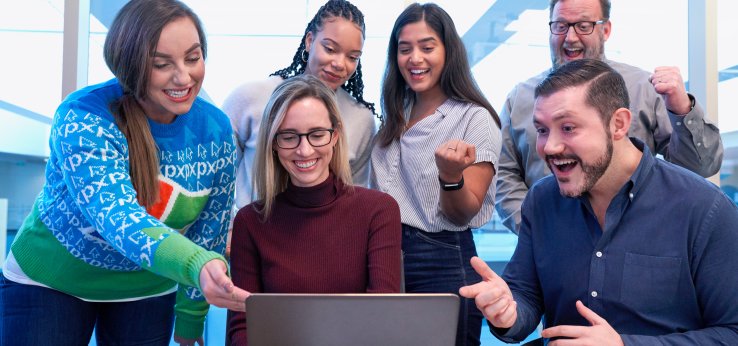 Vordergrund Laptop von hinten auf brauner Tischplatte. Hintergrund links 4 Frauen, rechts 2 Männer in einer Gruppe. Sie zeigen freudig und lachend auf den Laptop. (Bild: Windows / Unsplash)