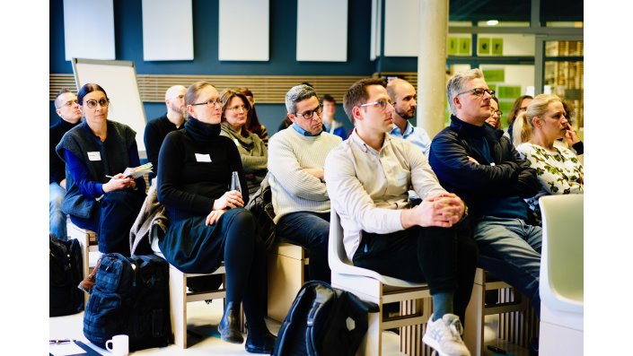 Die Teilnehmenden sitzen auf Stühlen und blicken zur Leinwand, die man im Bild nicht sieht.