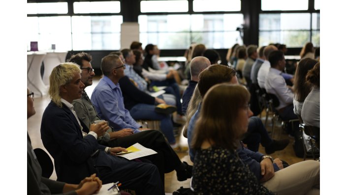 Das Publikum sitzt in Stuhlreihen seitlich zur Kamera und folgt der Veranstaltung.
