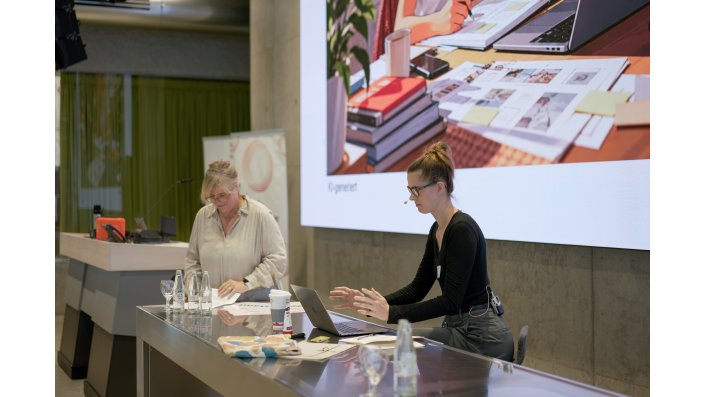 Foto: Zwei Frauen stehen an einem Tisch mit Laptops. Hinter ihnen ist eine Leinwand mit einer Präsentation.