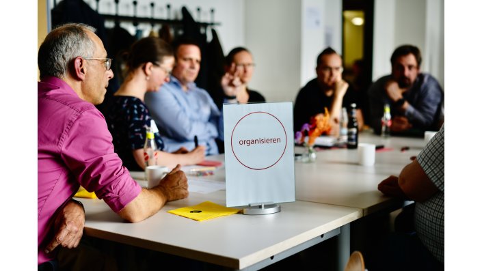 Sechs Männer und eine Frau sitzen an einem weißen Tisch mit Getränken, gelben Servietten und Kaffeetassen. Im Vordergrund des Bildes ist ein Schild auf dem das Wort ,,organisieren“ steht.