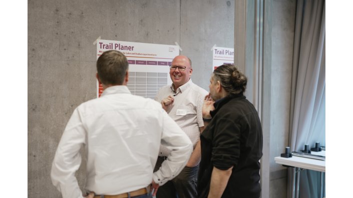 Foto: Drei Männer stehen vor zwei Plakaten mit der Überschrift ‚TraiL Planer‘ und unterhalten sich.