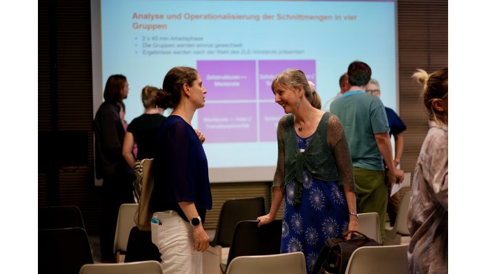 Mehrere Personen stehen vor einer Leinwand, auf die eine Präsentation projiziert ist, und unterhalten sich.