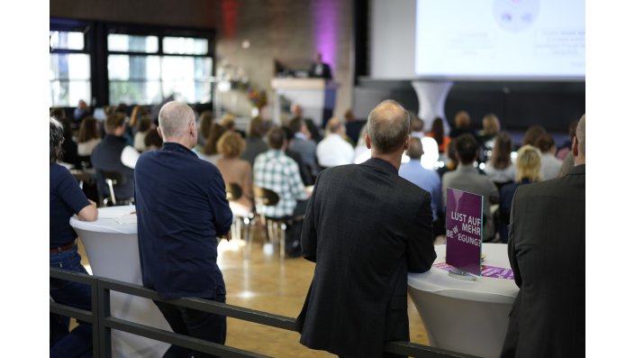  Mit dem Rücken zur Kamera stehen zwei Männer je an einen Stehtisch angelehnt und folgen der Veranstaltung.  