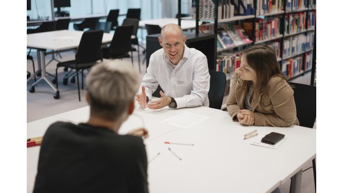 Foto: Zwei Frauen und ein Mann sitzen an einem Tisch in einem Raum mit Bücherregalen und unterhalten sich.