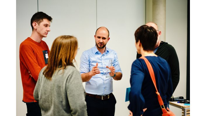 Ein Mann mit hellblauem Hemd steht in der Mitte und rechts und links stehen jeweils eine Frau und ein Mann. Die Gruppe diskutiert angeregt.