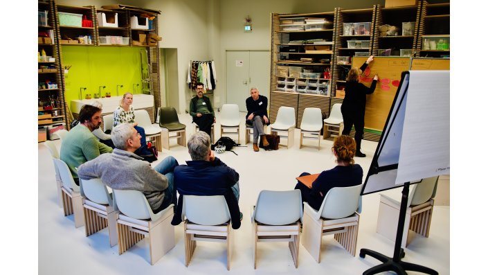 Im Stuhlkreis sitzen Prof. Dr. Axel Faßbender, Holger Müller, Emily Kobs, Timo van Treeck, Stefan Bente und weitere Teilnehmer*innen. [T(2.1]Rechts daneben steht Susanne Glaeser und hängt ein Post-It an eine Pinnwand.
