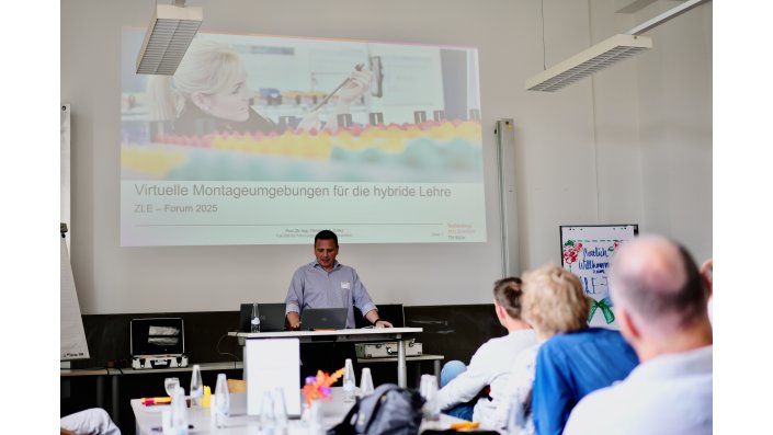 Prof. Dr. Christoph S. Zoller steht vor einem Rednerpult mit offenem Laptop. Hinter ihm ist eine Leinwand mit einer Power Point Präsentation, wo darauf steht ,,Virtuelle Montageumgebungen für die hybride Lehre ZLE – Forum 2025“. 