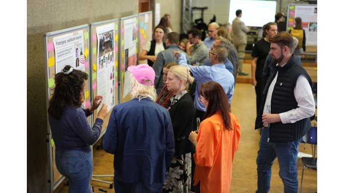 Vor einer Reihe an Pinnwänden stehen zahlreiche Personen und unterhalten sich in Kleingruppen. 