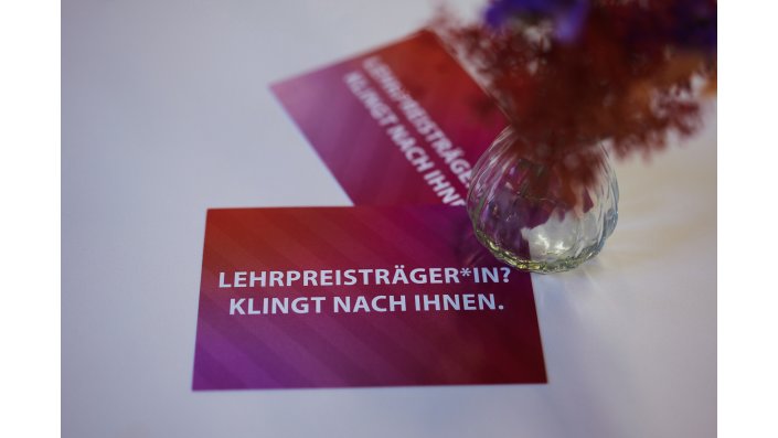 Zwei rote Postkarten mit weißer Schrift, die den Text 'LEHRPREISTRÄGER*IN? KLINGT NACH IHNEN.' tragen, daneben steht eine Vase mit Trockenblumen. 