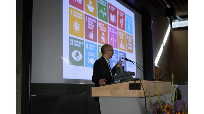 Prof. Dr. Matthias Barth steht an einem Rednerpult vor einer Leinwand und spricht zum Publikum. 