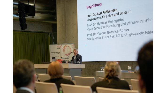 Foto: Ein Mann spricht an einem Rednerpult vor einem sitzenden Publikum. Hinter ihm ist eine Leinwand mit sichtbarem Begrüßungstext.