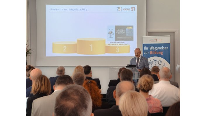 Unscharfes Publikum im Vordergrund. Im Hintergrund Herr Baetjer während der Laudatio in der Kategorie "Usability". Eine Präsentation mit Siegertreppchen ist geöffnet.