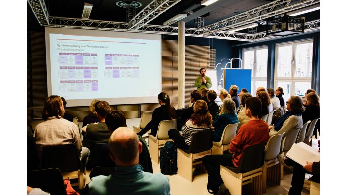 Im Vordergrund sitzen die Teilnehmenden und schauen auf die bespielte Leinwand. Rechts neben der Säule steht Timo van Treeck mit einem Mikrofon in der Hand. 